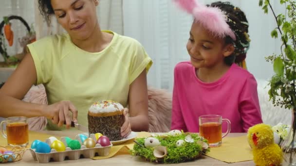 Mamma skär en påskkaka för att behandla sin dotter. Afroamerikansk kvinna och liten flicka sitter vid bordet i festligt inredda rum hemma. Glad påsk. Sakta i backarna. Närbild. — Stockvideo