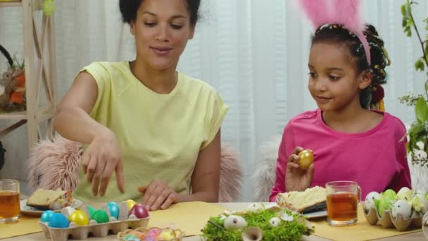 Mama i córka dobrze się bawią i rozbijają wielkanocne jajka. Afroamerykanka i dziewczynka siedzą przy stole w uroczyście urządzonym pokoju w domu. Szczęśliwego Wielkanocy. Zwolnij trochę. Zamknij się.. — Wideo stockowe