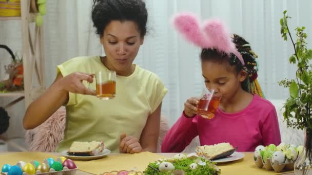 Mamma e figlia con divertenti orecchie da coniglio stanno bevendo il tè con la torta di Pasqua. Donna afroamericana e bambina sono seduti a tavola in una stanza decorata a festa a casa. Al rallentatore. Da vicino.. — Video Stock