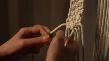 Yaratıcı bir atölyede bir kadın halat ustalığı yapıyor. Kadınların elleri makrome ipliklere düğüm atar ve yaratıcı bir dantel süsü yaratır. Boho tarzında makrome desenleri dokuyoruz. Kapatın. Yavaş çekim.