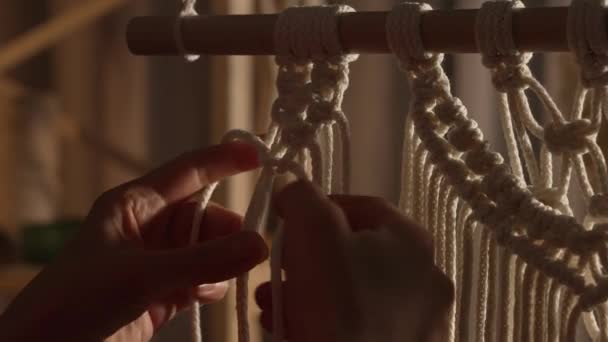 Vrouw weeft patroon van macrame draden met haar eigen handen voor decoratie of home decor. Womans handen knopen knopen aan touwen in een creatieve werkplaats in zacht licht. Handgemaakt. Sluit maar af. Langzame beweging. — Stockvideo