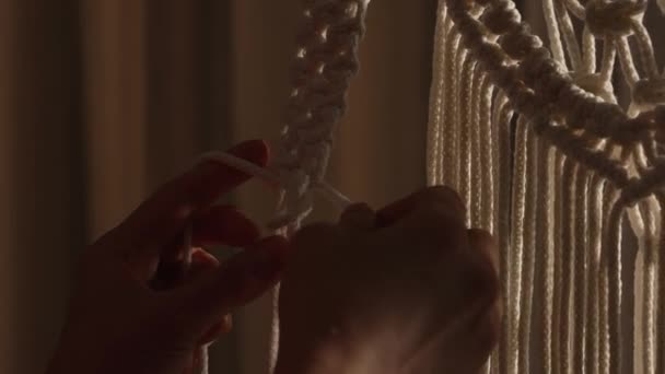 Woman weaves pattern of macrame threads with her own hands for decoration or home decor. Womans hands tying knots on ropes in a creative workshop in soft light. Handmade. Close up. Slow motion. — Stock Video