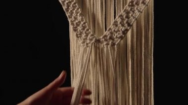 Yaratıcı atölyede kadınların elleri siyah arka planda bir makrome desen örer. Kadın dantel yapmak için pamuk ipliklere düğüm atıyor. Makromin temel düğümleri. İp ustası. Kapatın. Yavaş çekim.