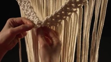 Doğal beyaz pamuk ipliklerinden makrome desenleri örüyorum. Kadınların elleri siyah arkaplanda dantel bir süs yaratarak düğüm atar. Atölyedeki yaratıcı süreç, tekstil el sanatları. Kapatın. Yavaş çekim.