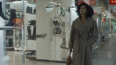 Mutlu Afrikalı Amerikalı kadın alışveriş merkezinde dolaşıyor ve markalı ayakkabı ve poşetlerle vitrinlere bakıyor. Modaya uygun ceketli bir kadın, büyük ve modern bir perakende mağazasında boş zaman geçiriyor. Yavaş çekim.