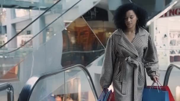 Стильная афроамериканка с красочными сумками в руках поднимается на эскалатор в торговом центре. Концепция шопинга, моды и розничной торговли. Женщина-покупатель гуляет среди витрин магазинов. Медленное движение. — стоковое видео