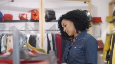 Kadın müşteri giyim mağazasından alışveriş yapıyor, askılarda asılı şık kıyafetler seçiyor. Güzel Afrikalı Amerikalı kadın yeni sezonluk koleksiyonundan bir şeyler seçiyor. Kapatın. Yavaş çekim.