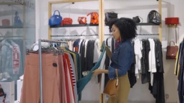 Kadın müşteri giyim mağazasından alışveriş yapıyor, askılarda asılı şık kıyafetler seçiyor. Güzel Afrikalı Amerikalı kadın yeni sezonluk koleksiyonundan bir şeyler seçiyor. Kapatın. Yavaş çekim.