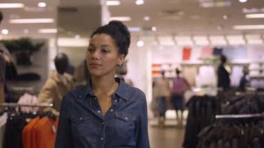 Güzel Afrikalı Amerikalı kadın moda giyim mağazasında askılarda asılı kıyafetleri seçiyor. Kadın müşteri bluz dener ve aynada kendine hayran kalır. Kapatın. Yavaş çekim.