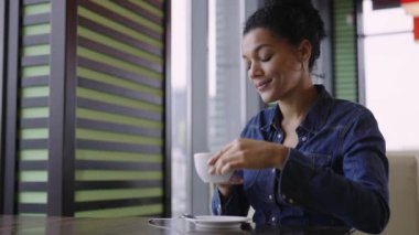Genç mutlu Afro-Amerikan kadın alışveriş merkezinin kafesindeki masalarda oturmuş aromatik kahvenin tadını çıkarıyor. Moda mağazasından başarılı bir alışveriş yapan bir kadın dinleniyor. Yaklaştın. Yavaş çekim.