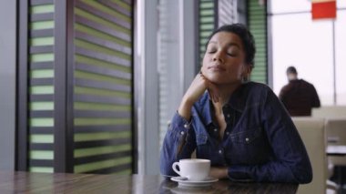 Yorgun ve genç Afrikalı Amerikalı kadın alışveriş merkezindeki kafede oturuyor. Bir fincan kahvenin üzerinde oturan üzgün bir kadının portresi. Kapatın. Yavaş çekim.