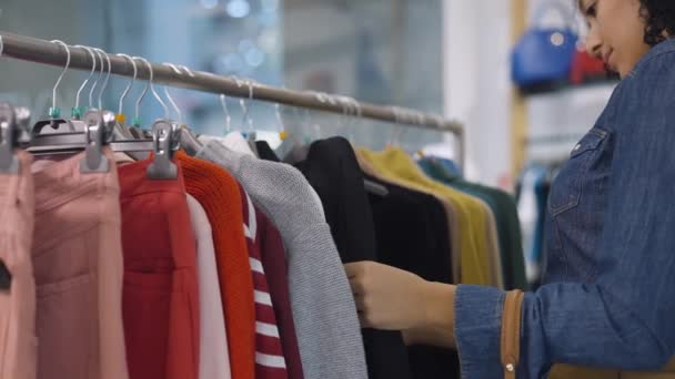 Cliente femenino hace compras en la tienda de ropa, elige ropa elegante colgando en perchas de rack. Hermosa mujer afroamericana elige cosas de la nueva colección estacional. De cerca. Movimiento lento. — Vídeos de Stock