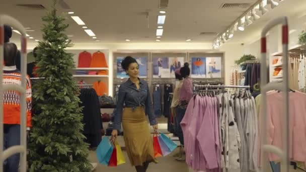 Mujer afroamericana feliz con bolsas de papel sale de la tienda de ropa de las mujeres en el centro comercial. Compradora femenina disfrutando de la compra en boutique de moda de marca en el centro comercial. De cerca. Movimiento lento. — Vídeos de Stock