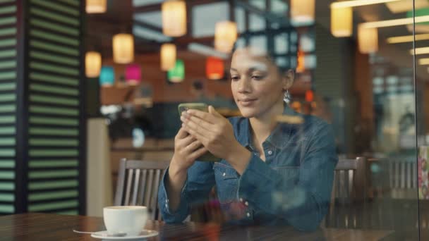Pohled přes skleněnou vitrínu mladé Afroameričanky textové zprávy na telefonu a těší kávu v obchoďáku kavárny. Žena odpočívá po úspěšném nákupu ve firemním obchodě s oblečením. Skoro. Zpomalený pohyb. — Stock video