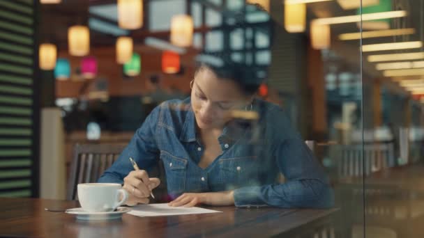 Blick durch ein Glasfenster einer jungen Afroamerikanerin, die verträumt an etwas denkt und dann auf einem Blatt Papier quietscht. Frau beim Kaffee in einem Einkaufszentrum-Café. Schließen. Zeitlupe. — Stockvideo