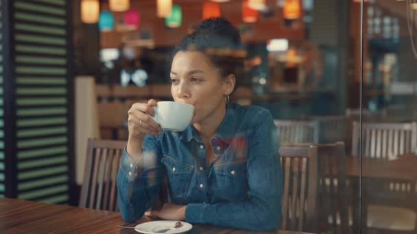 Vue à travers une vitrine en verre d'une jeune femme afro-américaine dégustant un café dans un centre commercial. Une femme se repose après avoir fait ses courses et fait signe à son ami de passer. Presque. Mouvement lent. — Video