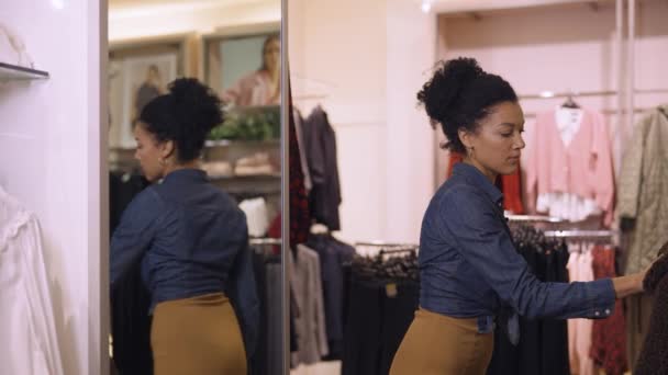 Mooie Afro-Amerikaanse vrouw die kleding kiest die hangt aan hangers in een trendy kledingwinkel. Vrouwelijke klant probeert een jas aan en bewondert zichzelf in de spiegel. Sluit maar af. Langzame beweging. — Stockvideo