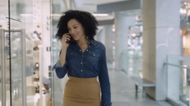 Gülümseyen Afrikalı Amerikalı kadın alışveriş merkezinde telefonla konuşuyor. Markette gezen şık bir kadın şık kıyafetler ve ayakkabılarla mağazanın camlarına bakıyor. Kapat..