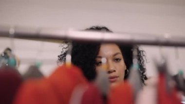 Güzel Afrikalı Amerikalı kadın moda bir giyim mağazasında askılarda asılı kıyafetleri seçiyor. Bir kadın alışveriş meraklısı, alışveriş merkezinden kıyafet alırken bir sürü elbisenin arasında koşar. Alışveriş konsepti