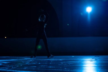 Genç bayan artistik patinajcı buz pistinde tek başına kadın paten koreografisi yapıyor. Yumuşak mavi ışık ve spot ışıklarının arka planına karşı buz arenasında pratik yapan bir kadın silueti..