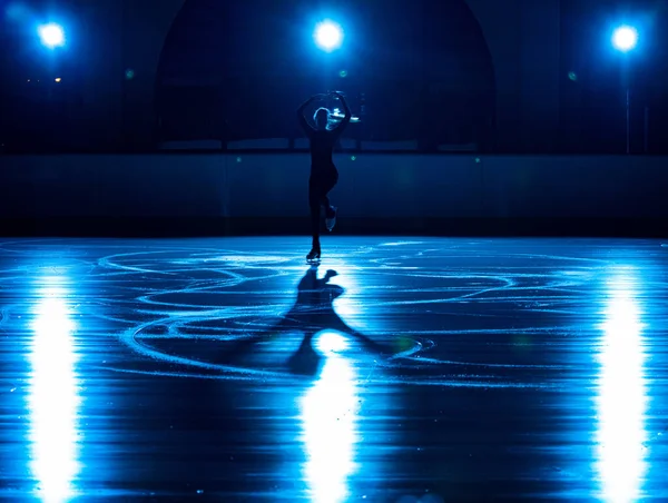 Young female figure skater is performing womans single skating choreography on ice rink. Woman practicing skills on ice arena against background of soft blue light and spotlights. Silhouette. — Stock Photo, Image