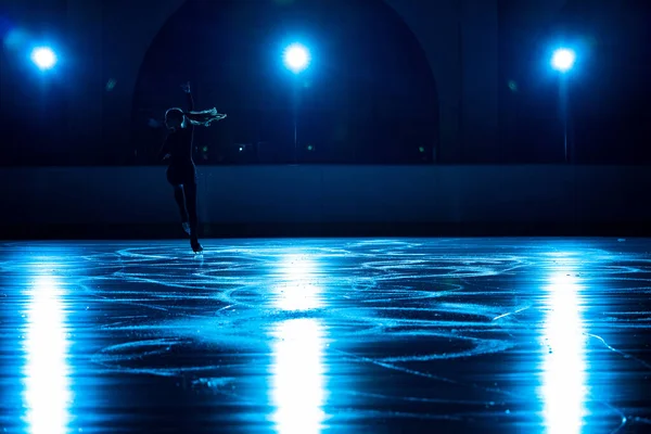 Jonge vrouwelijke kunstschaatsster voert enkele schaatschoreografie uit op de ijsbaan. Vrouw oefent vaardigheden op het ijs arena tegen de achtergrond van zacht blauw licht en spots. Silhouet. — Stockfoto