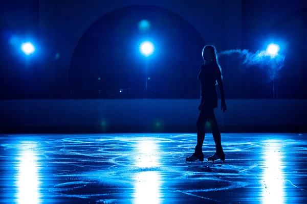 Ung kvinnlig konståkare utför kvinnlig singelskridskokoreografi på ishallen. Mörk siluett av kvinna öva färdigheter på isarena mot bakgrund av mjukt blått ljus och spotlights. — Stockfoto