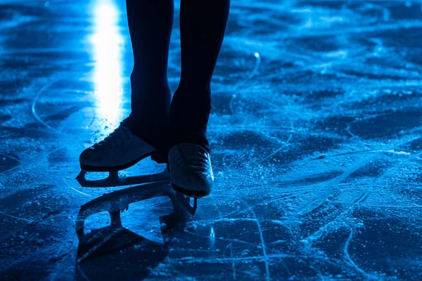 Detaljerad bild av kvinnors ben i vita konståkning skridskor på isarenan. Professionell idrottskvinna tränar på mörk ishall med blått ljus. Yta av is med repor och skridskobanor. Närbild. — Stockfoto