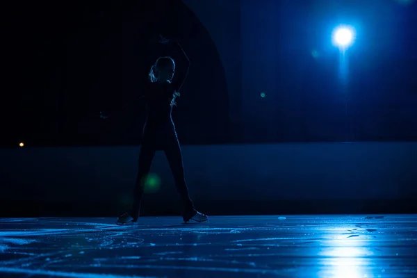 Jovem patinadora feminina está realizando coreografia de patinação única em pista de gelo. Silhueta escura de mulher praticando habilidades na arena de gelo contra fundo de luz azul suave e holofotes. — Fotografia de Stock