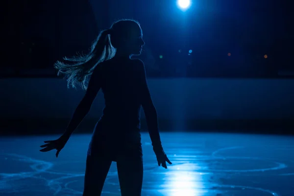 Mladá krasobruslařka provádí ženy choreografii na kluzišti. Silueta ženy cvičit dovednosti na ledové aréně na pozadí modrého světla a reflektorů. Zavřít. — Stock fotografie