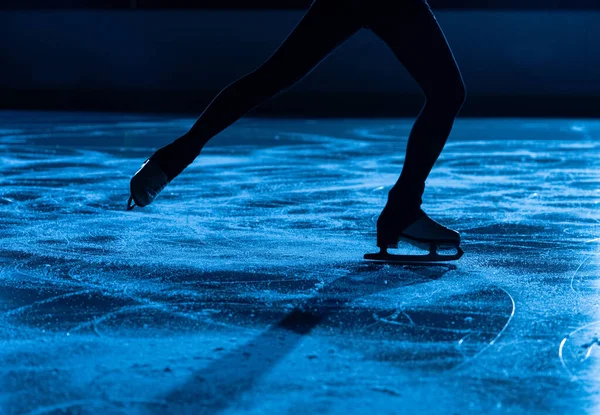 Ciemna sylwetka kobiecych nóg w łyżwach figurowych ślizgających się po lodowej arenie. Młoda kobieta trenuje na ciemnym lodowisku z niebieskim światłem. Błyszcząca gładka powierzchnia lodu z zadrapaniami od łyżew. Zamknij się.. — Zdjęcie stockowe