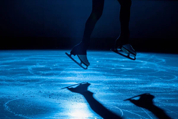 The female athlete in figure skating at the moment of the jump. Womens legs in white skates for figure skating. Practicing skating skills on a dark ice arena with blue light. Close up.