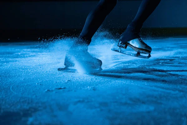 Szczegółowy strzał kobiecych nóg w białe łyżwy figurowe na zimnej arenie lodowej w ciemności z niebieskim światłem. Kobieta ślizga się po lodzie, rozpryskując cząsteczki lodu do kamery. Zamknij się.. — Zdjęcie stockowe