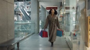 DNYPRO, UKRAINE - 1 Aralık 2021: Zarif Afro-Amerikan kadın alışveriş merkezlerinde kağıt alışveriş torbalarıyla yürüyor. Kadınlar markalı giysilerle mağazada başarılı alışveriş yapmaktan zevk alırlar. Yavaş çekim.