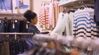 DNYPRO, UKRAINE - 1 Aralık 2021: Güzel Afrikalı Amerikalı kadın moda giyim mağazasında askılarda asılı kıyafetleri seçiyor. Bayan müşteri giyim mağazasından alışveriş yapıyor. Kapatın. Yavaş çekim.