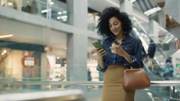 DNYPRO, UKRAINE - 1 ДЕКАБРЯ 2021: Счастливая африканская американка пишет смс на iphone и пьет кофе, стоя в торговом центре. Женщина отдыхает и общается во время покупок в магазинах одежды. Закрыть. — стоковое видео