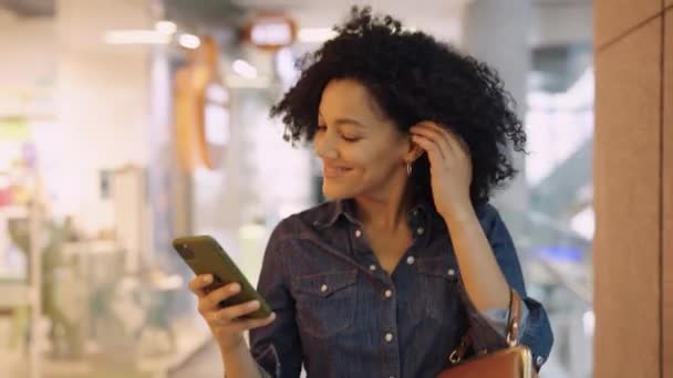 华盛顿，英国- 2021年12月1日：快乐的非洲裔美国女人在购物中心的橱窗里散步，用iphone发短信。购物和时尚的概念。有品牌服装的大型现代商店。靠近点 — 图库视频影像