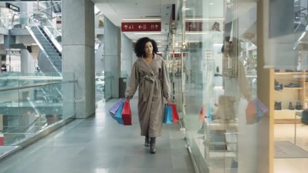 DNYPRO, UKRAINE - DECEMBER 1, 2021: Stijlvolle Afro-Amerikaanse vrouw loopt door het winkelcentrum met heldere papieren boodschappentassen. Een vrouw op zoek showcase met merkkleding in een winkel. Langzame beweging. — Stockvideo