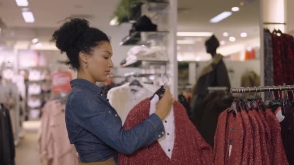 华盛顿，英国- 2021年12月1日：非洲裔美国女人选择挂在服装店衣架上的衣服。女顾客试穿夹克，并在镜子中欣赏自己。靠近点慢动作. — 图库视频影像