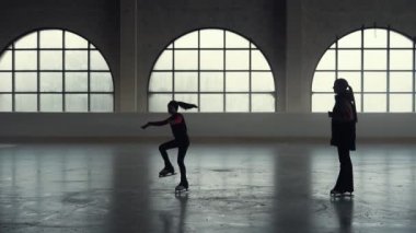 Artistik patinaj eğitimi. Küçük kız artistik patinajcı tek bacakla paten kayıyor. Kadın antrenör, karanlık bir buz arenasında yumuşak ışıkla bir öğrenciye öğretiyor. Koyu renk siluetler. Yavaş çekim.