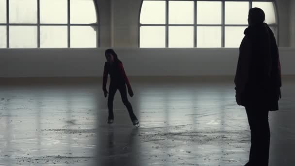 Treinadora ensina patinação artística a uma adolescente. Pequena atleta treina, praticando saltos e habilidades de giro em uma arena de gelo escuro com luz suave. Sílhuetas escuras. Movimento lento. — Vídeo de Stock