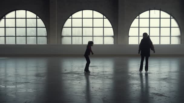 Žena trenér ukazuje své dívce studentské prvky krasobruslení choreografie. Holčička krasobruslařka bruslí a trénuje své jednonohé rotace dovednosti na temné ledové aréně. Siluety. Zpomalený pohyb. — Stock video