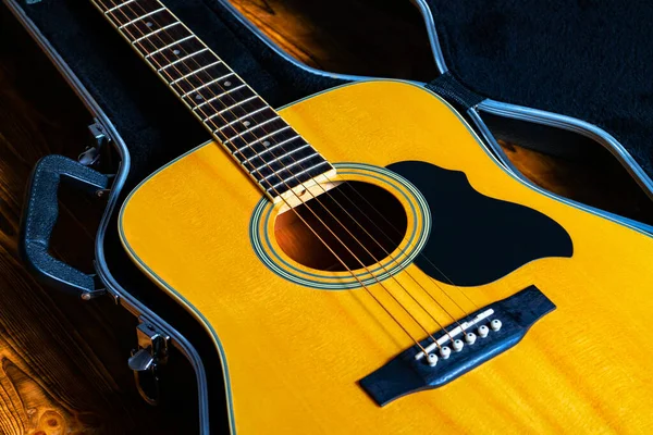 Tahta sarısı akustik gitar sert bir çantada duruyor. Siyah bir gardırop bagajında telli müzik aleti. Akustik gitarın bir kısmı fretboard, ses deliği ve tellerle kapanır..