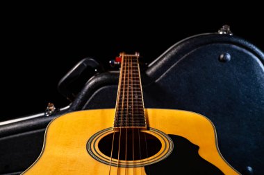Tahta sarısı akustik gitar sert bir çantada duruyor. Siyah bir gardırop bagajında telli müzik aleti. Akustik gitarın bir kısmı fretboard, ses deliği ve tellerle kapanır..
