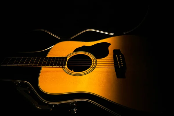 Sárga akusztikus gitár fekve egy kemény tok a sötétben, fekete háttérrel. Fa húros műszer, fénysugárral megvilágítva. Gitár fedél közelről. Rock, folklór, country zene. — Stock Fotó