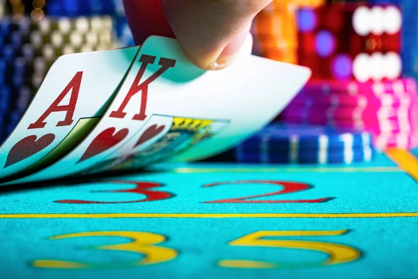 プレーヤーはカジノの青いゲームテーブルに横たわっているトランプを持ち上げます。王とエースを明らかにする男の手。チップとサイコロのぼやけた背景にポーカーカードを閉じます。. — ストック写真