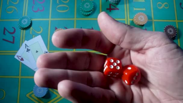Male hand shakes red dice in slow motion against background of green roulette table in a casino. Man gambler, gambling, craps, poker. Casino chips and cards are laid out on the gaming table close up. — ストック動画