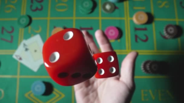 Male hand tossing up red dice in slow motion against background of green roulette table in casino. Man gambler, gambling, craps, poker. Casino chips and cards are laid out on gaming table close up. — ストック動画