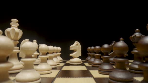 Camera pans over chessboard with chess pieces placed and focuses on white horse. White and brown wooden chess set on squared board on black background. Wooden figures for playing chess close up. — стоковое видео