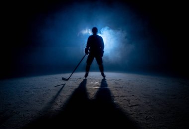 Dark silhouette of a male hockey player in a uniform, helmet and skates with a stick on the ice arena with smoke and blue back light. Sportsman posing at the stadium ice rink. Winter sports.